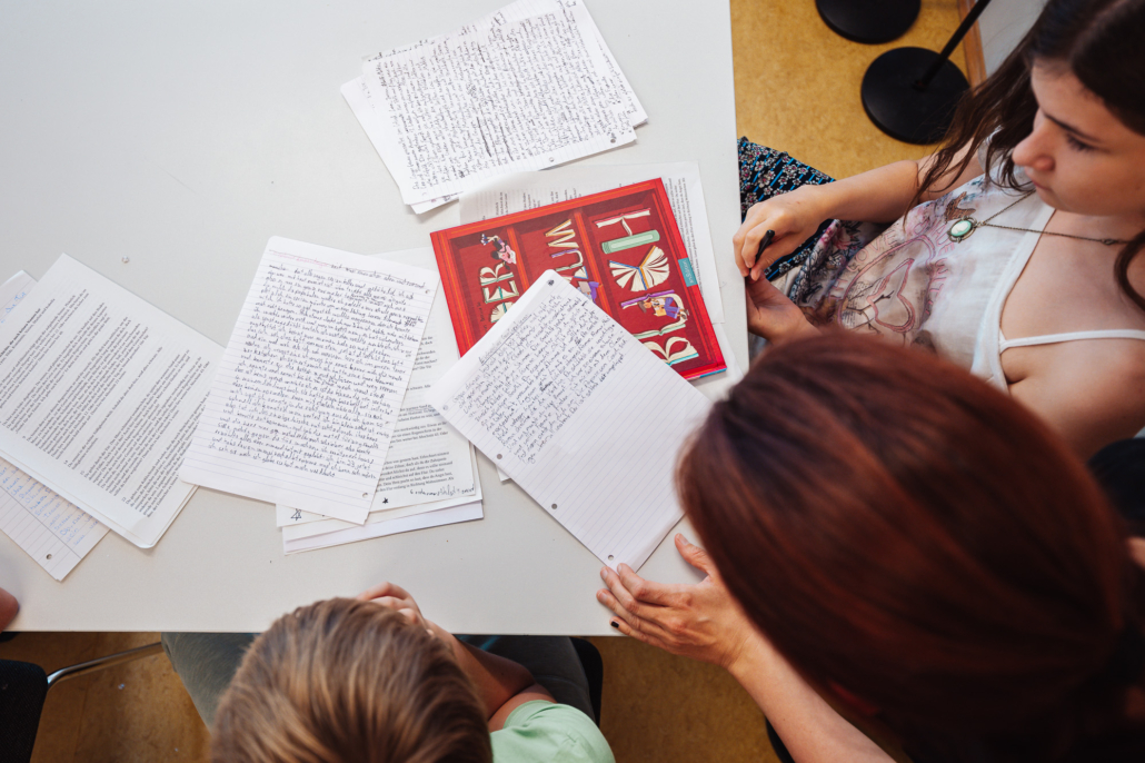 Handgeschriebene und Computer geschriebene Blätter liegen sortiert aufeinaner auf einem Tisch, darunter ein rotes Deckblatt mit Buchstaben.